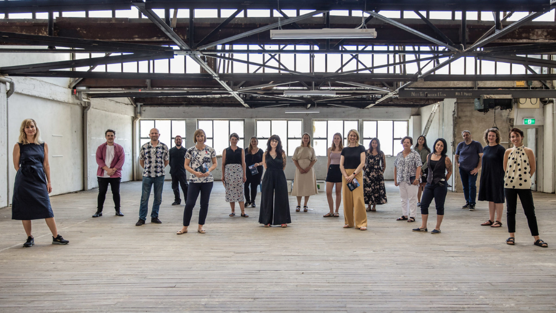 The Village Project Team stands in a line facing camera in an empty warehouse