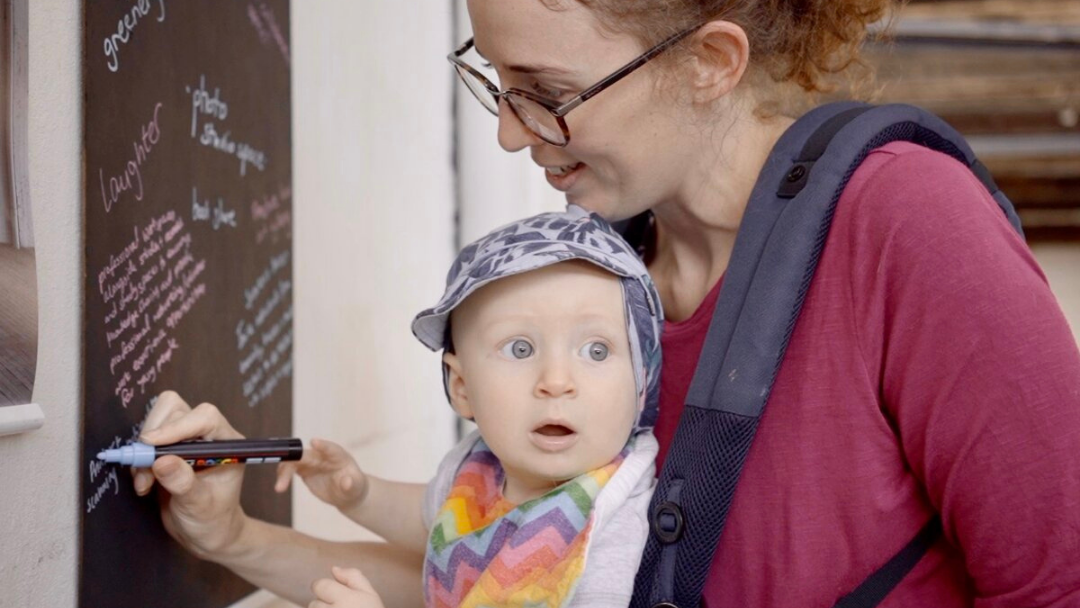 Parent writes on a blackboard holding a baby