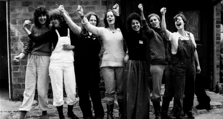 a line of smiling women face camera with their arms raised in strength and solidarity